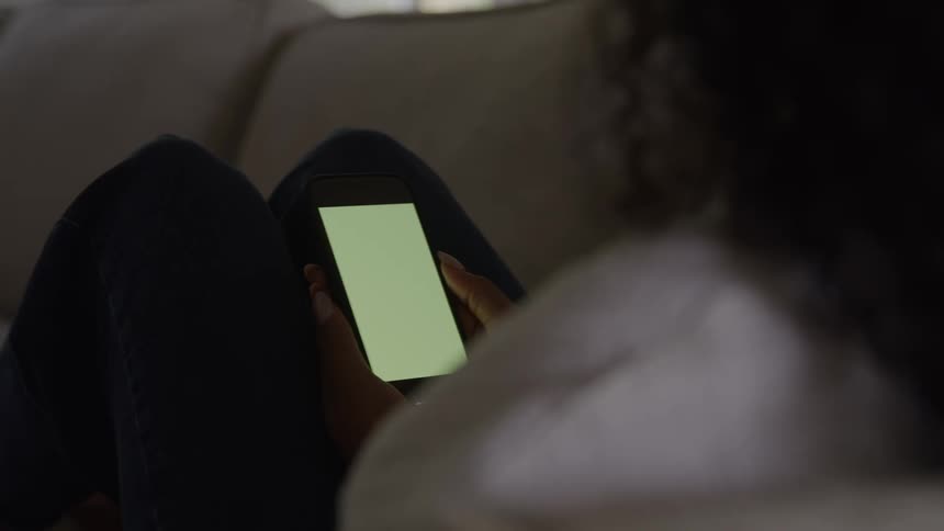 A woman is seen in a dark intimate indoor setting resting her legs while intently holding and interacting with a smartphone