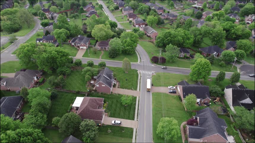 An aerial view shows a school bus driving through a lush green suburban neighborhood