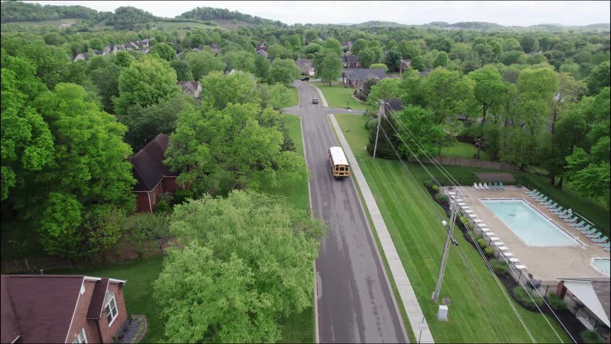 A school bus drives along a suburban street at midday with buildings and trees lining both sides of the road