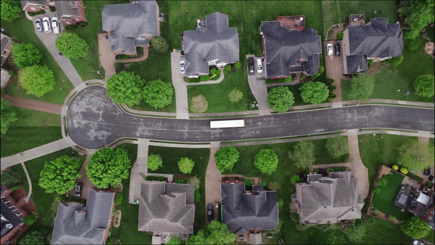 A t suburban neighborhood street showcasing a grid of large houses surrounded by bright green lawns and mature trees as a lone white bus drives down the curved asphalt road progressing from the center toward the bottom left of the frame