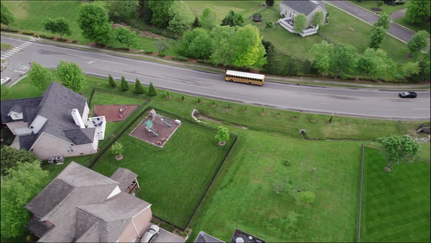 A school bus drives along a suburban street at midday with buildings and trees lining both sides of the road