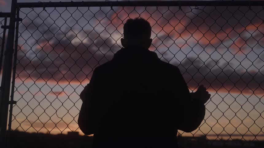 A man stands leaning against a wire fence at sunset gazing thoughtfully over the horizon