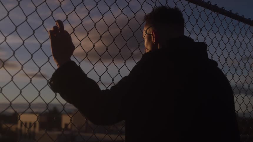 A man holds onto a chain link fence while staring through it
