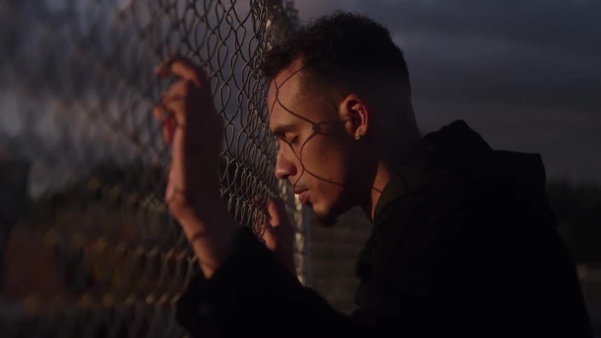 A young man leans against a chainlink fence with his eyes closed the warm light of sunset casting gridlike shadows across his contemplative face