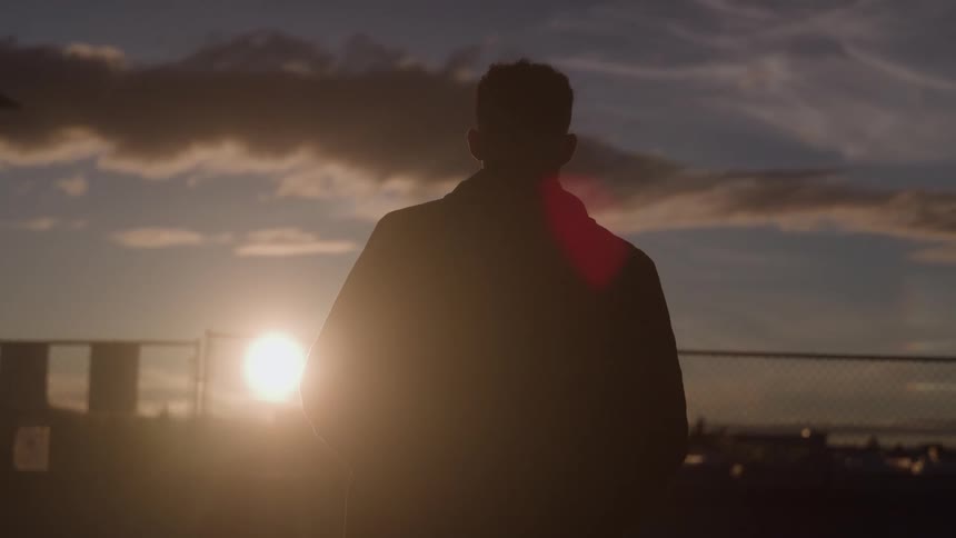 A man is silhouetted from behind wearing a jacket and walking against a dramatic sunset sky with warm light behind dark clouds and a chainlink fence in the distance