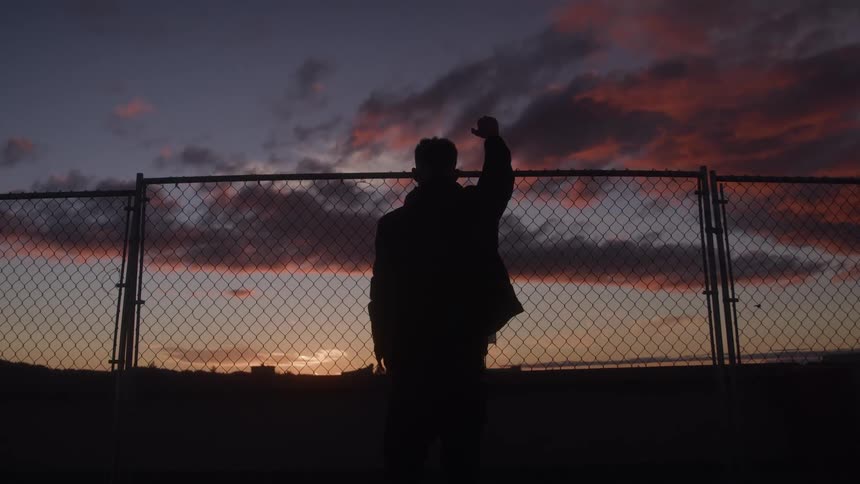 A man is dramatically silhouetted from behind wearing a jacket and raising a triumphant fist above a chainlink fence against a vivid sunset sky filled with orange and purple clouds