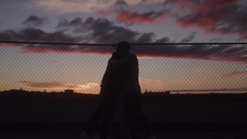 Silhouette of two men walking toward each other next to a chain link fence then shake hands and embrace