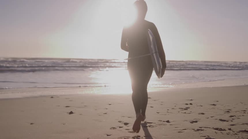 A surfer viewed from behind and completely silhouetted against the bright sun runs along a sandy beach toward the ocean the individual is holding a surfboard under one arm and appears ready to enter the water for a surf session