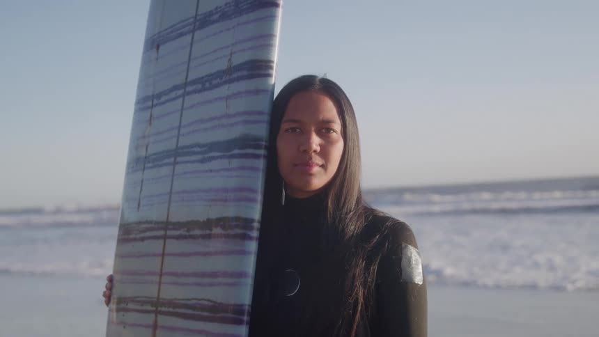 Woman standing on the beach holding her surfboard as waves roll into shore