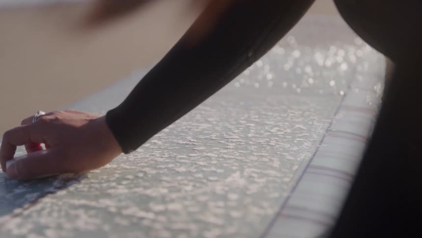 A person in a wetsuit applies a bar of wax in circular motions to the textured surface of a surfboard in the bright sunlight