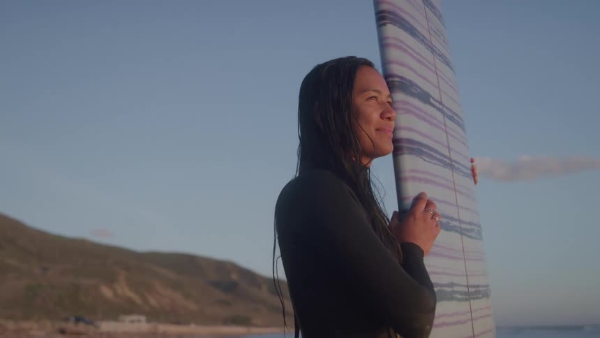 Female surfer smiles while standing on the beach and holding a surfboard