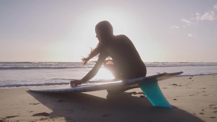 A surfer kneels on the beach silhouetted against the bright sun setting over the ocean as they apply wax to their surfboard the surfboard with its bright blue fin facing the viewer rests on the sand near the gently washing waves