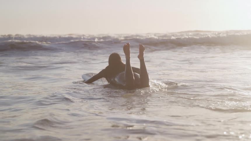 Woman paddling on her surfboard toward the waves at sunrise