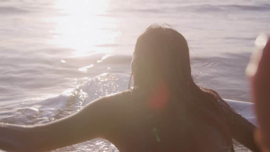Woman lying on surfboard in the ocean at sunset facing the glowing horizon
