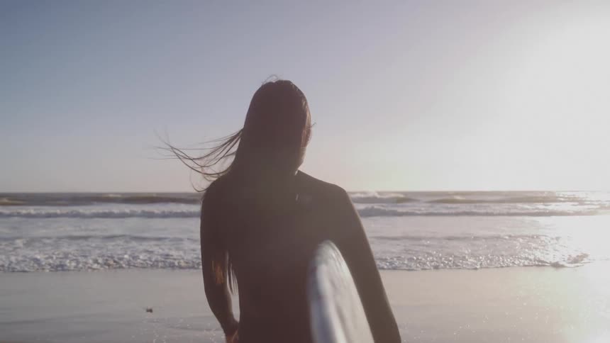 A surfer seen from behind and silhouetted against a bright sun walks towards the ocean waves while holding a surfboard the sun flares brightly over the water creating a warm golden atmosphere at the beach
