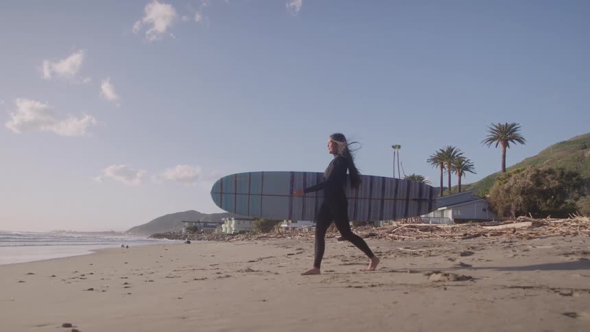 A woman in a wetsuit walks along a sandy beach carrying her surfboard with the ocean and a coastal neighborhood in the background