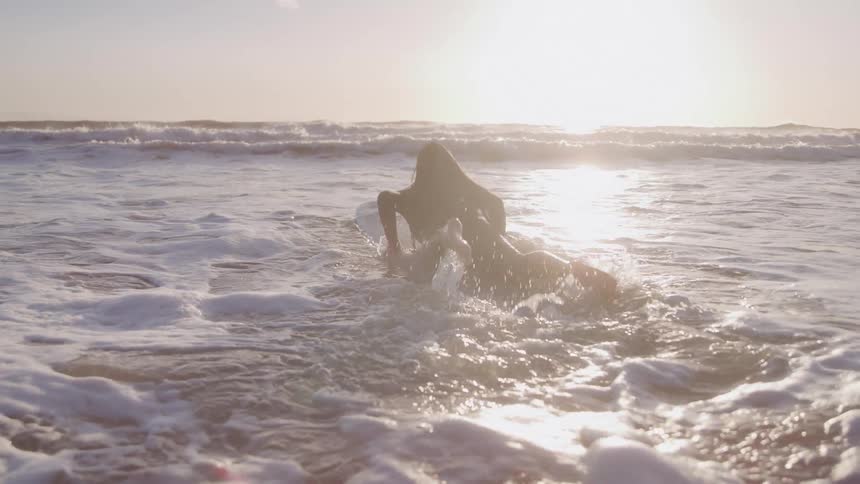 A person with long hair seen from behind wades into the ocean with a surfboard during sunset they crouch down allowing the waves to wash over them and the board before paddling out