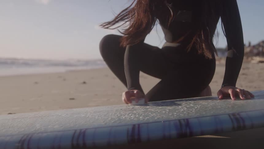 The video shows a closeup of a person dressed in a black wetsuit sitting on a sandy beach near the ocean they are applying wax to the deck of a surfboard with a bar of wax