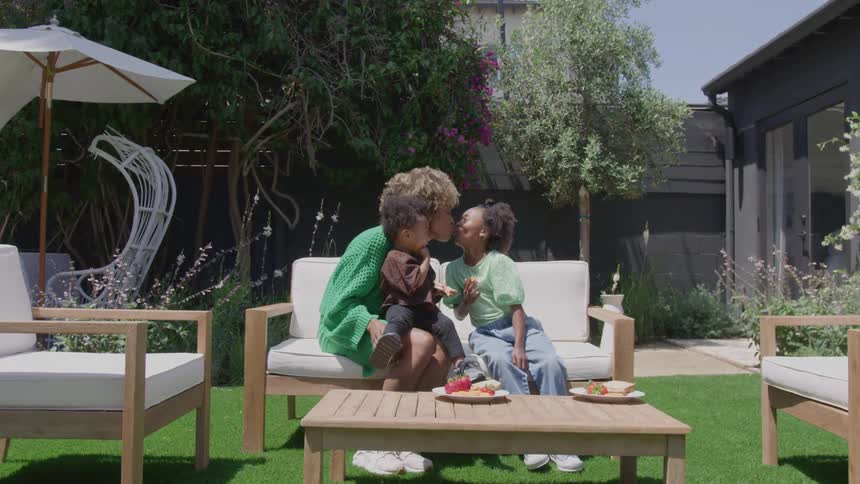 A mother sits at midday under the sun with an infant on her lap eating fruit from the table in front of her while gently kissing her young daughter beside her
