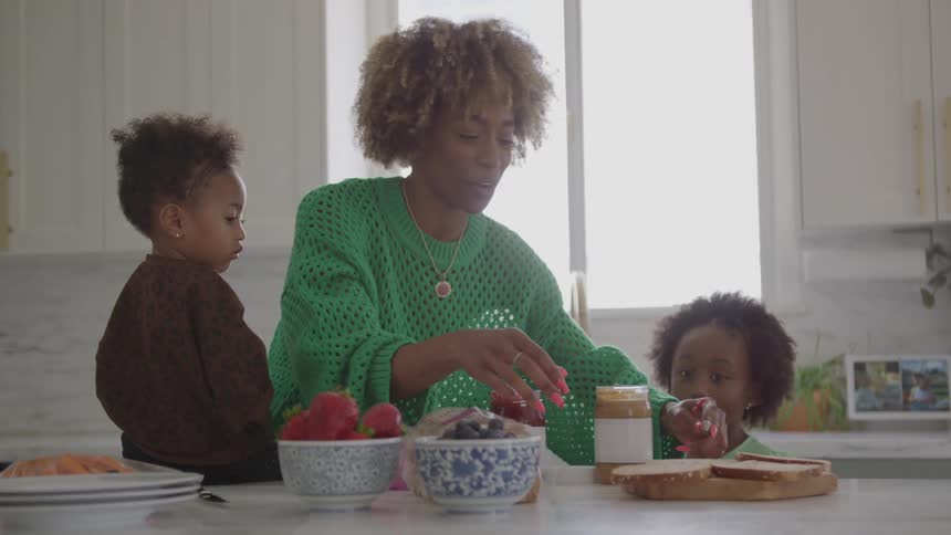 In a kitchen a mother wearing a green sweater stands over a countertop smiling and happily preparing a sandwich with a knife while her two young children one on either side of her watch intently