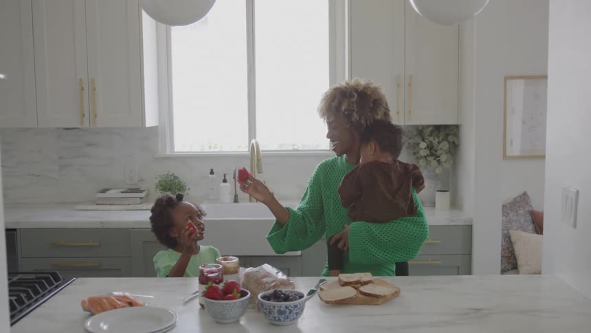 A mother holding an infant interacts with her older daughter while standing in a bright kitchen the mother is smiling and giving a strawberry to the daughter who is seated at the counter where ingredients for a meal are laid out