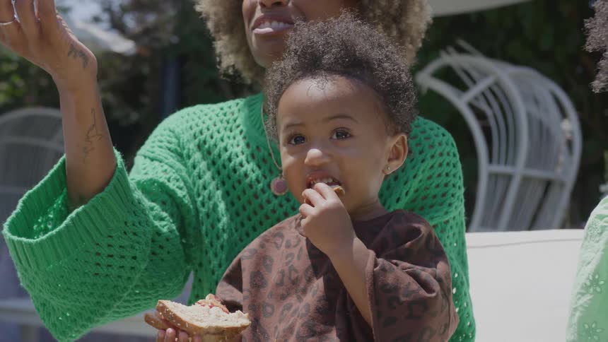A girl eats a sandwich while she sits with an adult female