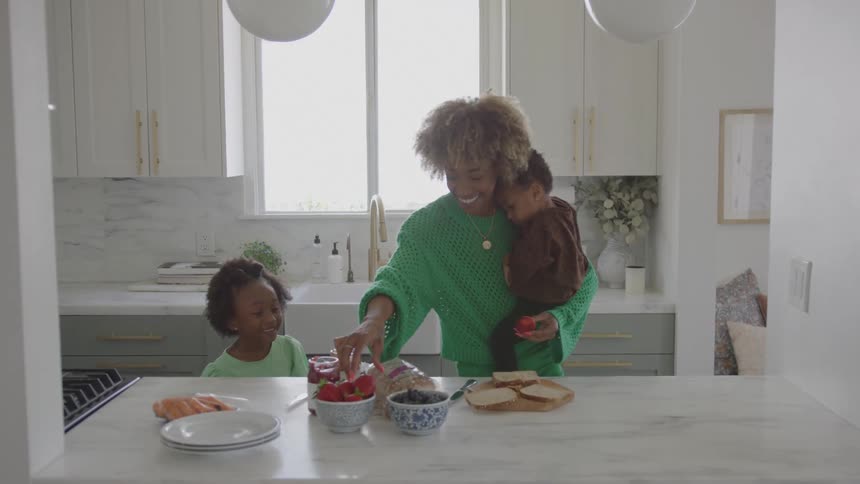 Mother holding her toddler prepares a meal as she and her daughter smile while standing in the kitchen during the daytime