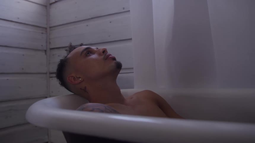 A young man with curly hair and a tattoo is captured in a contemplative moment reclining topless in a white bathtub against a backdrop of rustic wooden paneling looking thoughtfully upward towards the diffused light