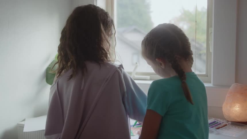 Two young girls stand sidebyside at a desk looking out a bright window one with braids drawing on paper as the other watches