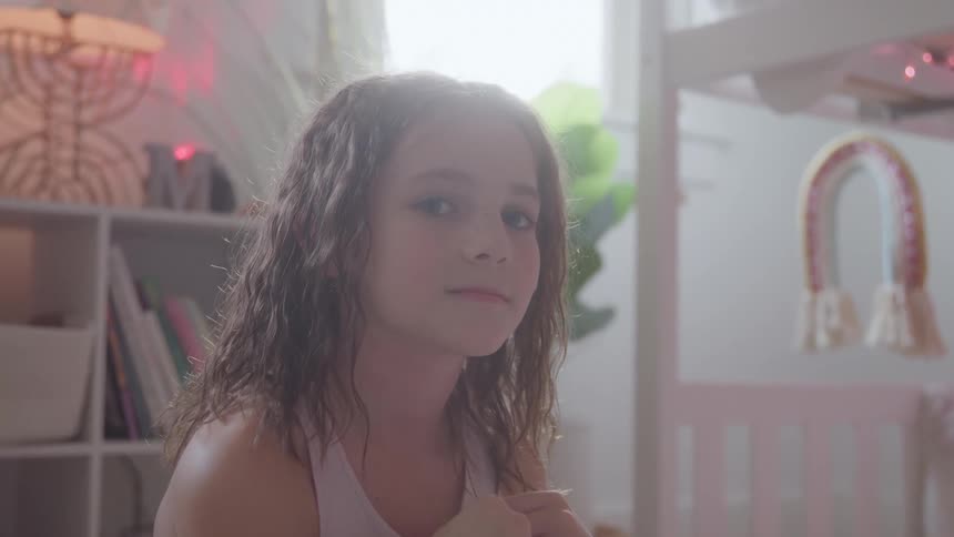 The video features a young girl with long brown hair wearing a white top looking slightly offcamera with a neutral expression she is in a bright room with shelves and colorful decorative lights in the background