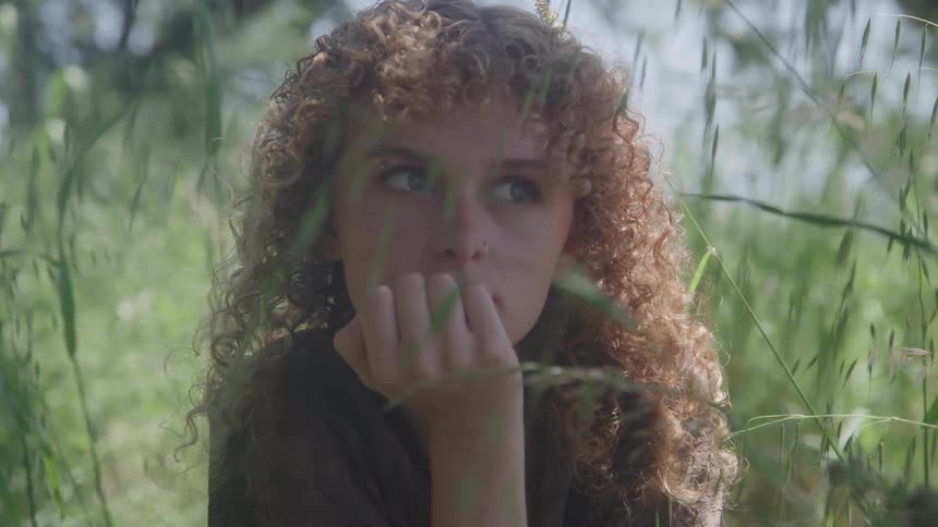 Girl holds her hand to her face as plants and vegetation moves in the breeze