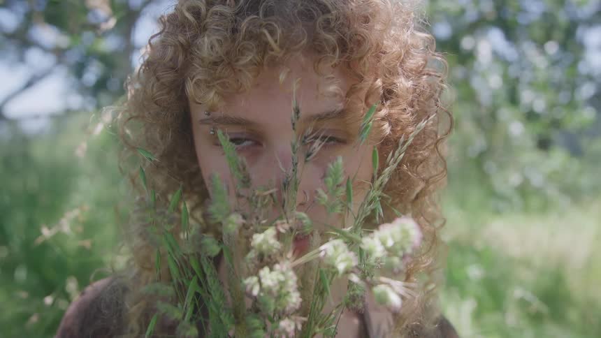 A woman with striking green eyes and bright tightly coiled blonde hair gazes intently over a handful of green grasses and delicate white wildflowers in a sundappled overgrown field