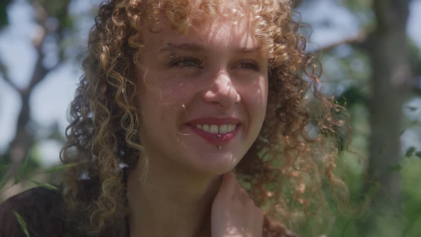 A closeup shot captures a smiling woman with curly hair looking at the camera as dappled sunlight filters through the trees behind her