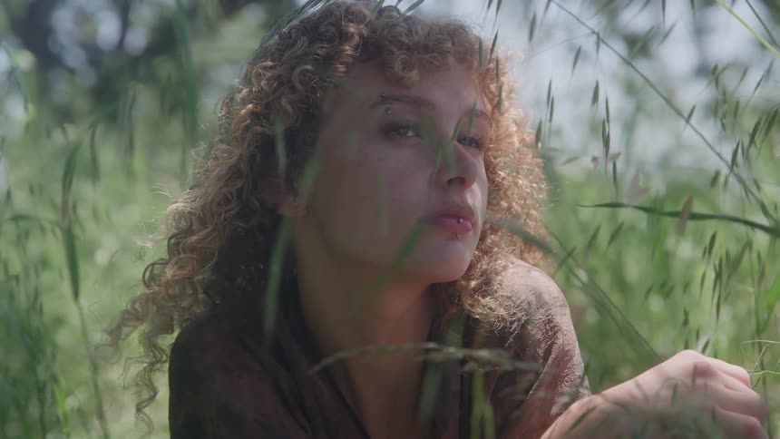 A young woman with tight curly hair sits in a sundappled field of tall green grass she looks upwards with a contemplative expression as the light catches her face