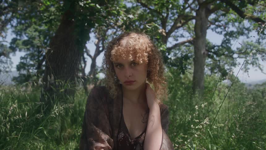 A young woman with curly blonde hair looks up with a serene expression while sitting in a sunlit grassy field
