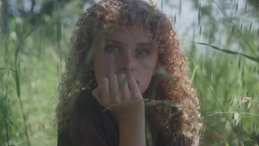 A young woman with long curly brown hair rests in the woods at midday surrounded by tall grasses her right arm gently placed against her cheek