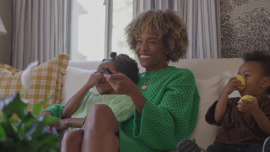 A mother sits on a couch at midday in the living room holding a remote with excitement one daughter laughs while lying against her and the toddler sits beside her eating an orange fruit