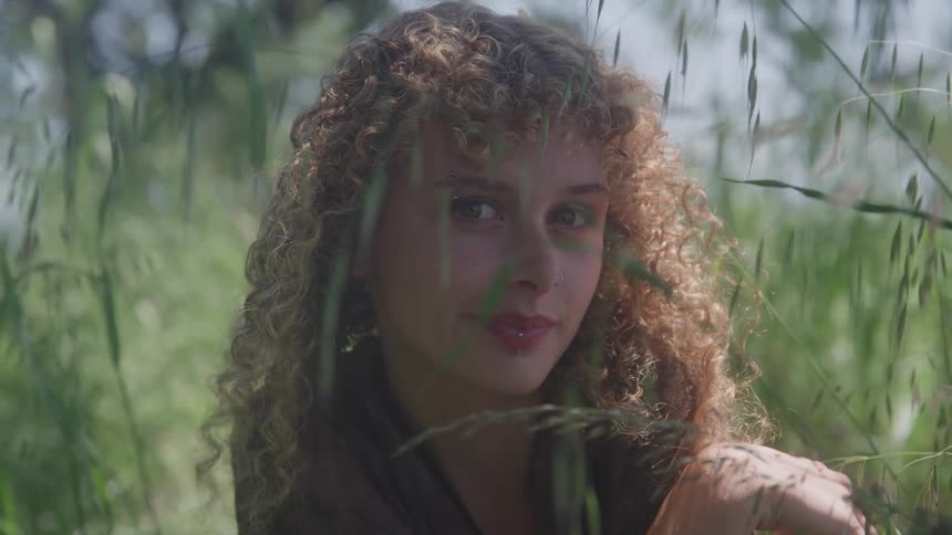 A woman with curly hair sits among tall blades of grass turning her gaze from the side to look directly at the camera with a soft smile