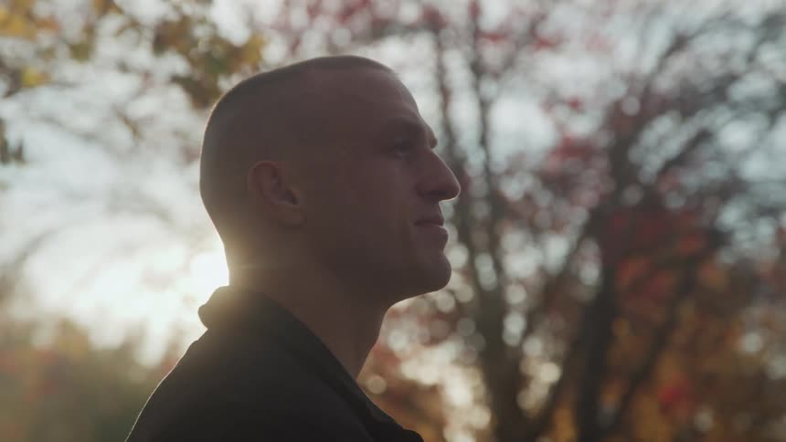 A man with a buzzed haircut is seen in profile against a bright blurry background of autumn trees slowly tilting his head to look up toward the sky