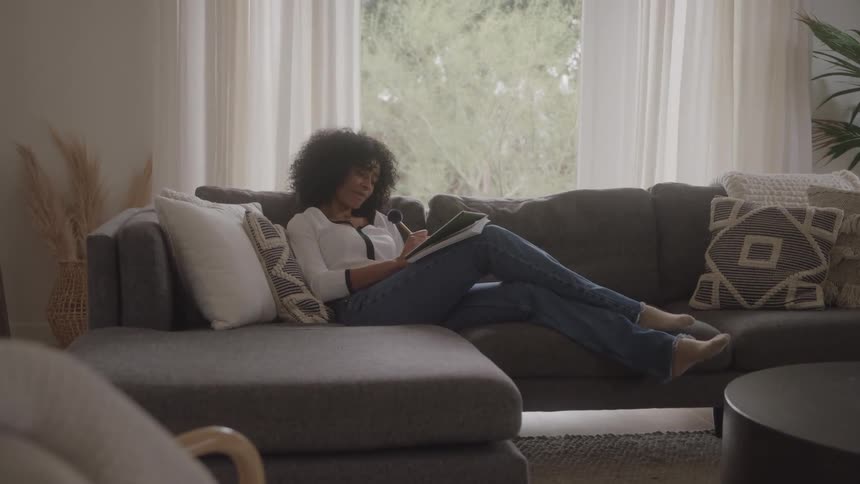 Woman sitting on her living room couch smiles while writing in a notebook