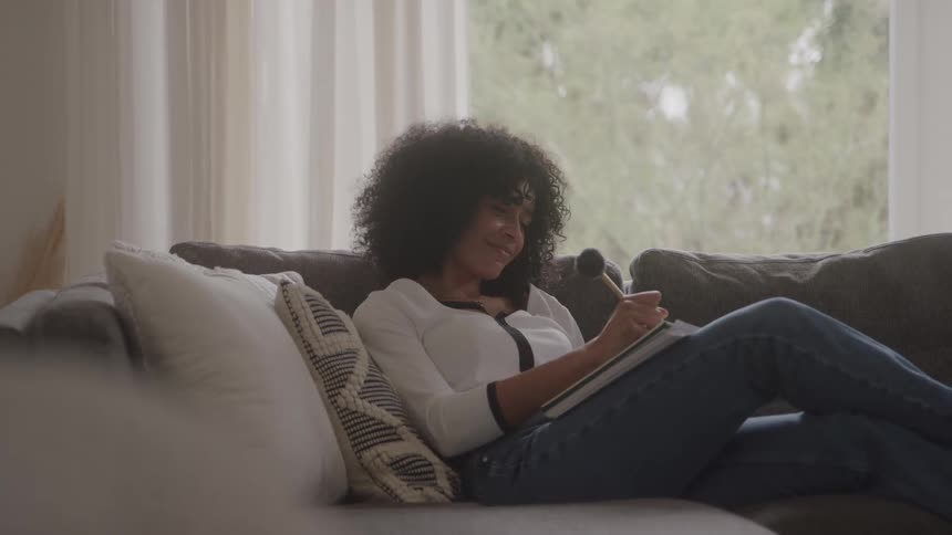 Woman sitting on her living room couch smiles as she writes in a book