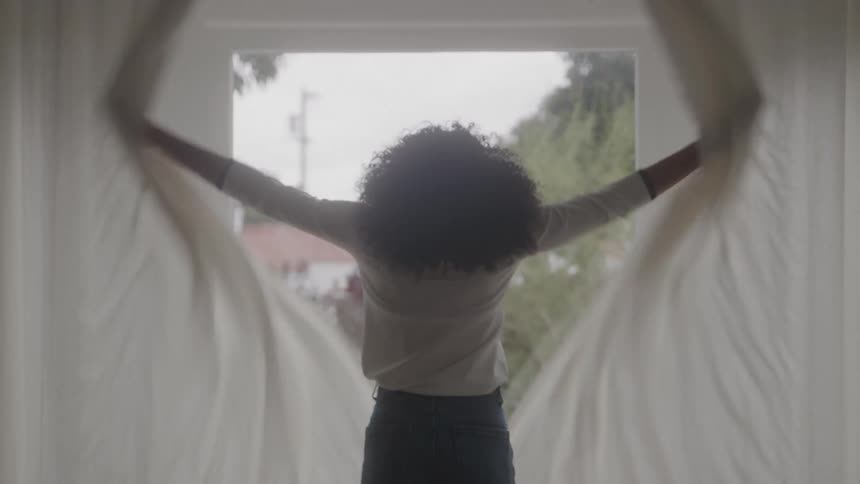 Woman walks across the room to the window and opens the curtains