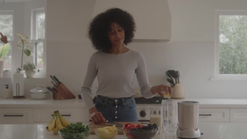 A smiling woman with dark curly hair is standing in a bright modern kitchen cutting a piece of fruit on a wooden cutting board with various bowls of smoothie ingredients like berries kale and bananas laid out on the counter