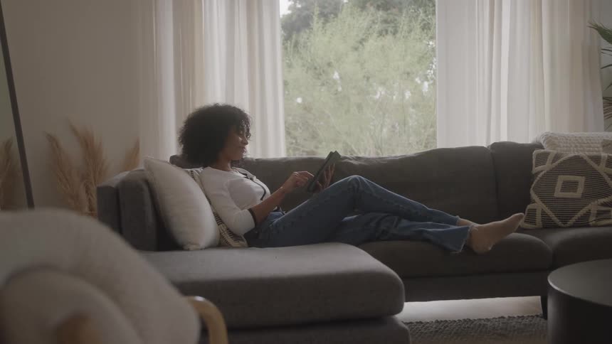 Person relaxing on gray sofa with tablet in sunlit room featuring cozy decor