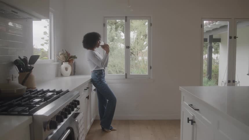 A woman with curly hair stands in a bright modern kitchen leaning against the counter while looking out a window she holds a cup close to her mouth appearing to be enjoying a warm drink and a quiet moment