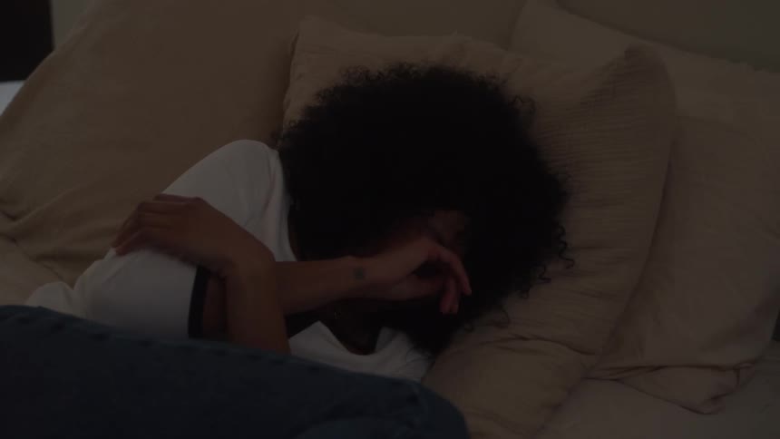 A black female with dark voluminous curly hair is pictured lying on her side in a dimly lit bed wearing a white shirt and blue jeans appearing comforted as she cries while hugging herself against a large textured beige pillow