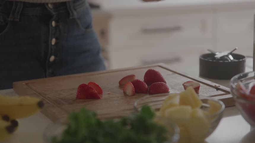 A woman is standing in a kitchen using a silver knife to slice fresh red strawberries on a wooden cutting board surrounded by bowls of other fruits