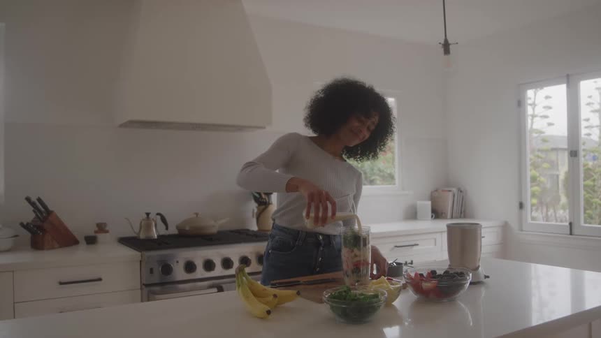 On a woman with dark curly hair and dressed in casual attire stands in her spacious brightly lit white kitchen focused on the counter where she is actively preparing a smoothie by carefully adding liquid on a tall blender jar