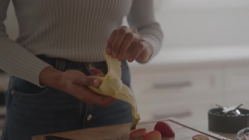 A person wearing a light sweater and jeans is in a kitchen peeling a banana over a wooden cutting board the board already holds some cut fruit including strawberries and pineapple suggesting they are preparing a meal or snack