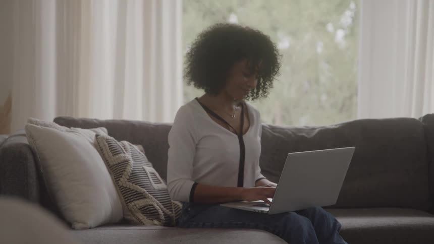 A smiling woman with dark voluminous curly hair is seated comfortably on a plush gray sofa in a brightly lit room typing actively on a silver laptop while wearing a white longsleeved shirt and blue jeans